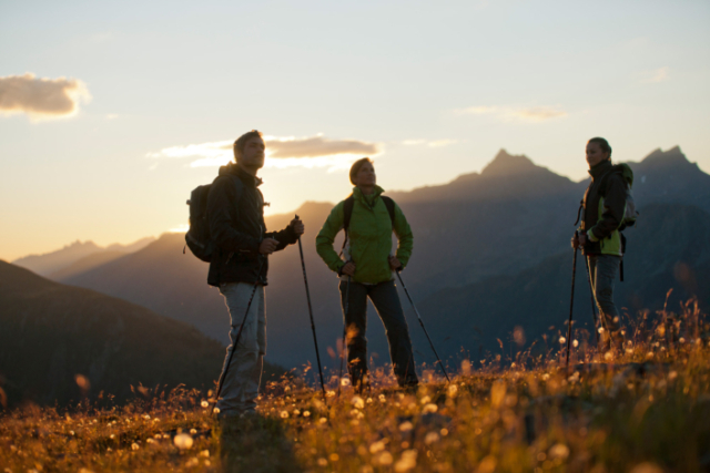 Ein Mann und zwei Frauen stehen mit ihrer Wanderausrüstung auf einem Berg und schauen in die Sonne.